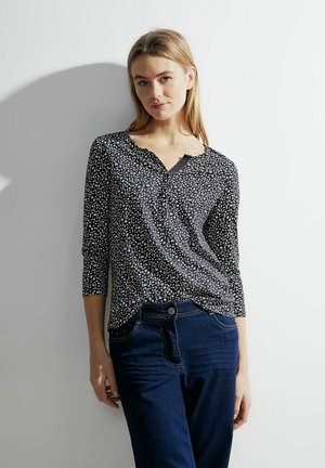 Schwarz-weiß gemusterte Bluse mit V-Ausschnitt, dreiviertel Ärmeln und abgerundetem Saum, kombiniert mit dunkelblauen Jeans.