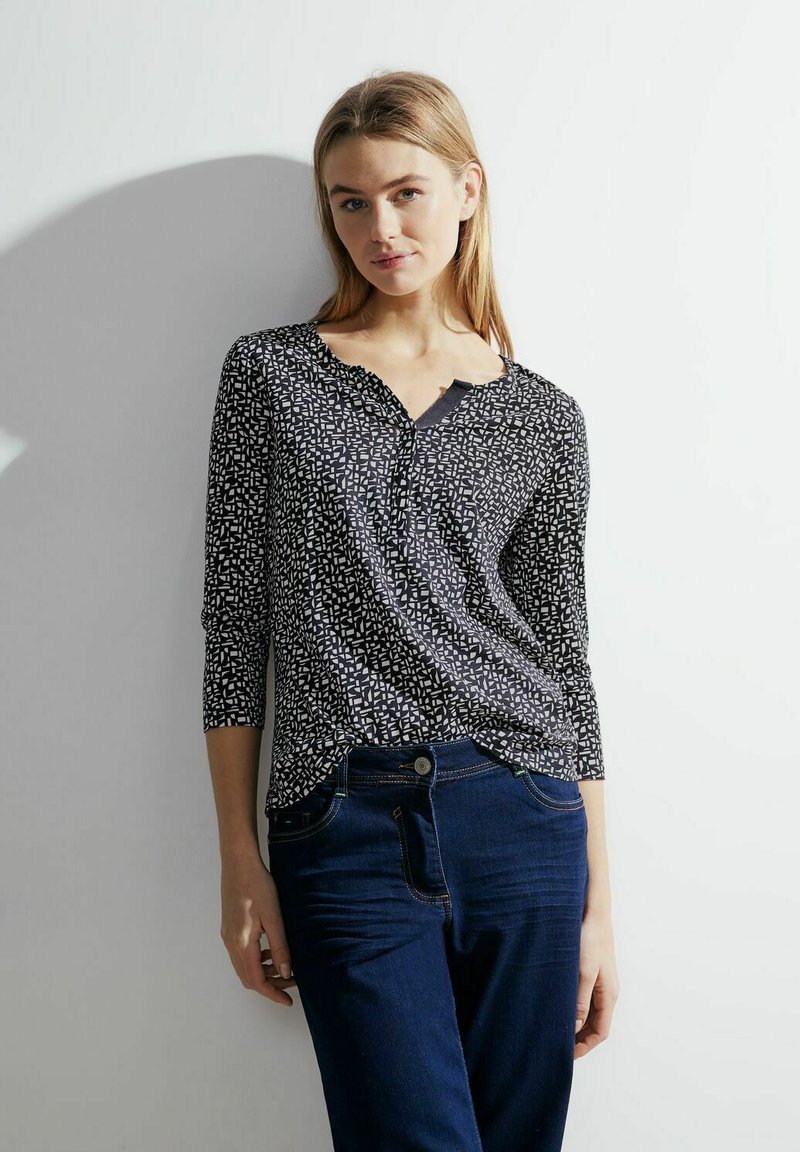 Czarna i biała bluzka w wzory z dekoltem w kształcie litery V, rękawami o długości 3/4 i zaokrąglonym dołem, połączona z ciemnoniebieskimi dżinsami.