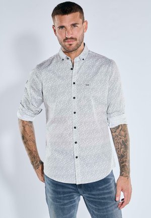 Weißes Hemd mit schwarz-blumigem Muster, langärmlig hochgekrempelt, und schwarzen Knöpfen. Kombiniert mit blauen Jeans.