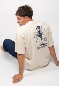 Béžové bavlněné tričko s krátkým rukávem. Na zadní části je grafika ženy, která drží nápoj, s textem "Pátek večer" a "Na zdraví, Drahá."