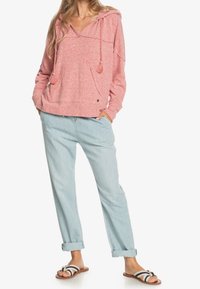 Sudadera rosa con capucha, acabado texturizado y bolsillos delanteros; combinada con pantalones azul claro con puños doblados y sandalias a rayas.