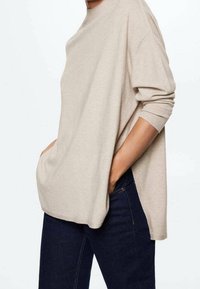 Pull beige en maille avec un col rond, épaules larges tombantes et fentes latérales, associé à un jean bleu foncé, mettant en valeur une silhouette décontractée.