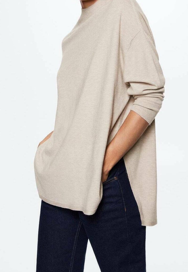 Pull beige en maille avec un col rond, épaules larges tombantes et fentes latérales, associé à un jean bleu foncé, mettant en valeur une silhouette décontractée.