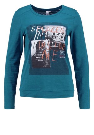 Chemise manches longues bleu sarcelle avec graphique en média mixte et texte en blanc, orange et noir sur le devant, incluant la phrase "SPACES INSIDE ME".