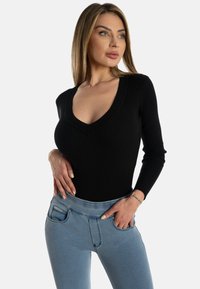 Vaaleahiuksinen nainen, jolla on päällään musta pitkähihaisen V-kaula-aukon paita ja vaaleansiniset farkut, poseeraa kädet lanteilla valkoista taustaa vasten.