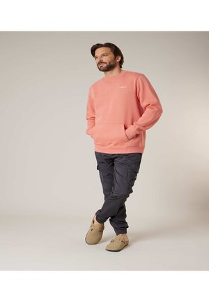 Homme portant un sweat corail, un pantalon cargo foncé et des chaussures beige sans lacets, debout les mains dans les poches et les jambes croisées.