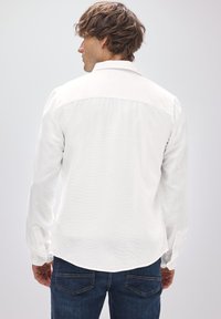 Weißes Langarmhemd mit Kentkragen, strukturiertem Stoff, abgerundetem Saum und Manschetten mit Knopfverschluss, kombiniert mit blauen Jeans.