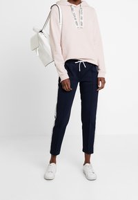 Sudadera con capucha de color rosa claro con tirantes con logo, pantalones deportivos azul marino con rayas laterales blancas y zapatillas blancas. Los accesorios incluyen un bolso de hombro blanco.