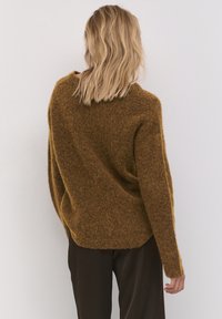 Kobieta z blond włosami do ramion nosi luźny, brązowy, puszysty sweter oraz ciemne spodnie, stojąc na tle jasnym, plecami do obiektywu.