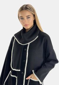 Cappotto nero con un ampio colletto, dettagli in cucitura bianca, chiusura frontale con bottoni e tasche oversize con un motivo testurizzato.