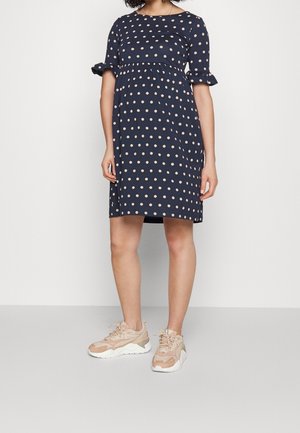 Frau trägt ein dunkelblaues knieumspieltes Kleid mit beigen Polka-Dots und beigen klobigen Sneakers, steht vor einer schlichten Wand.