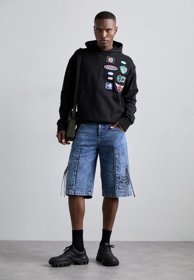 Μαύρη hoodie με διάφορα patches, μπλε τζιν σορτσάκια με φερμουάρ στις τσέπες, μαύρα αθλητικά παπούτσια και μαύρες κάλτσες. Απλός φόντο.