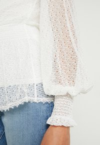 Top bianco in pizzo con maniche a sbuffo, dotato di motivi intricati e polsini arricciati, abbinato a denim blu. Texture e dettagli di design ben visibili.