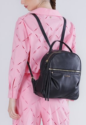 Donna che indossa un completo rosa traforato e porta uno zaino in pelle nera con cerniere dorate e una tasca frontale.