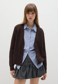 Cardigan de punto marrón con un corte relajado, llevado sobre una camisa de botones a rayas azul claro y blanca, y una falda gris carbón con cordón.