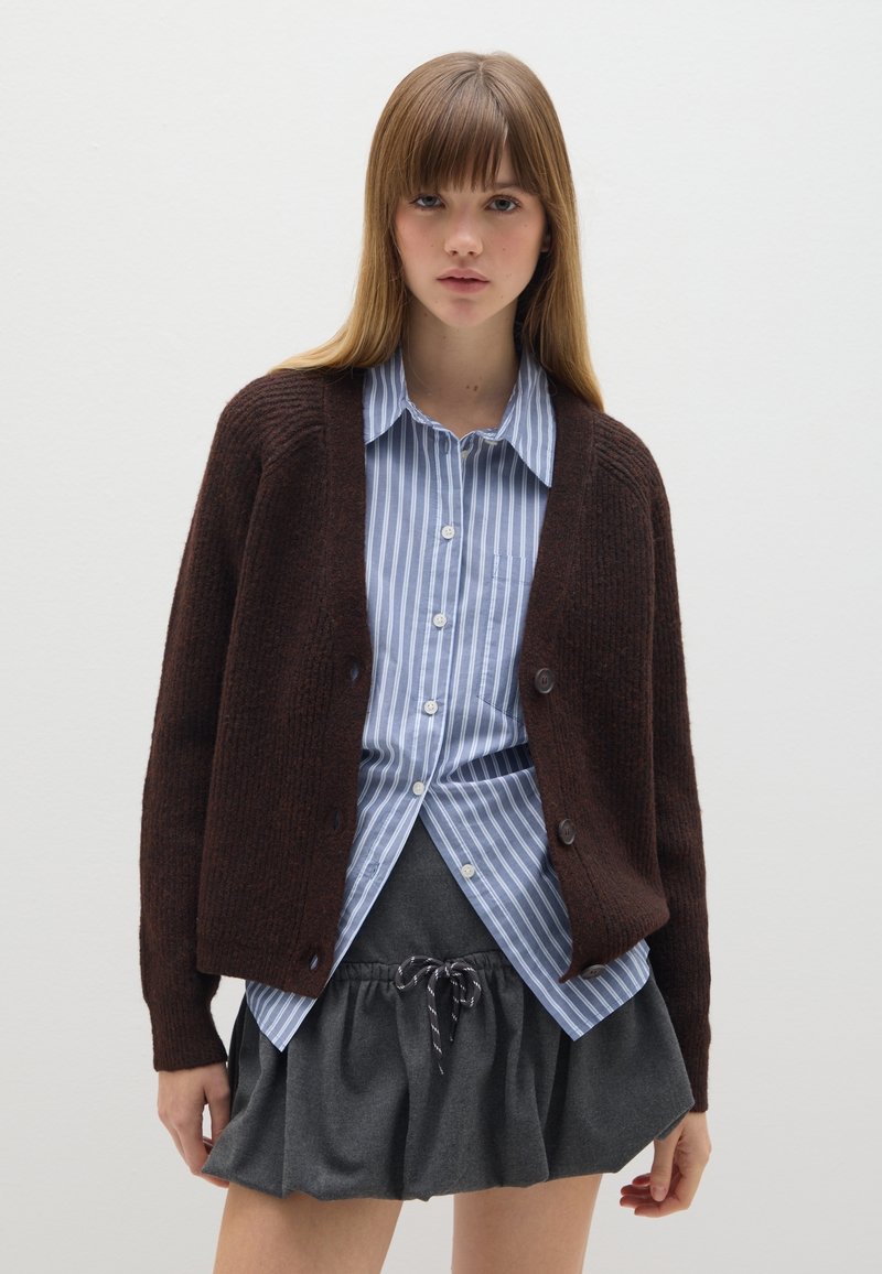 Cardigan de punto marrón con un corte relajado, llevado sobre una camisa de botones a rayas azul claro y blanca, y una falda gris carbón con cordón.