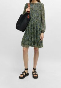 Blumenmuster, knielanges Kleid mit langen Ärmeln, in grünen und blauen Farbtönen, kombiniert mit einer schwarzen Ledertasche und schwarzen Riemchensandalen.