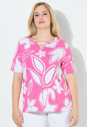 Frau trägt ein pinkfarbenes Kurzarmoberteil mit großen weißen Blumenmustern und weiße Hosen, steht vor einem weißen Hintergrund.