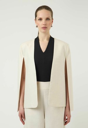Mujer con el pelo peinado hacia atrás, vestida con una capa-blazer crema sobre una camiseta negra de cuello en V y pantalones crema a juego, de pie delante de un fondo claro y liso.