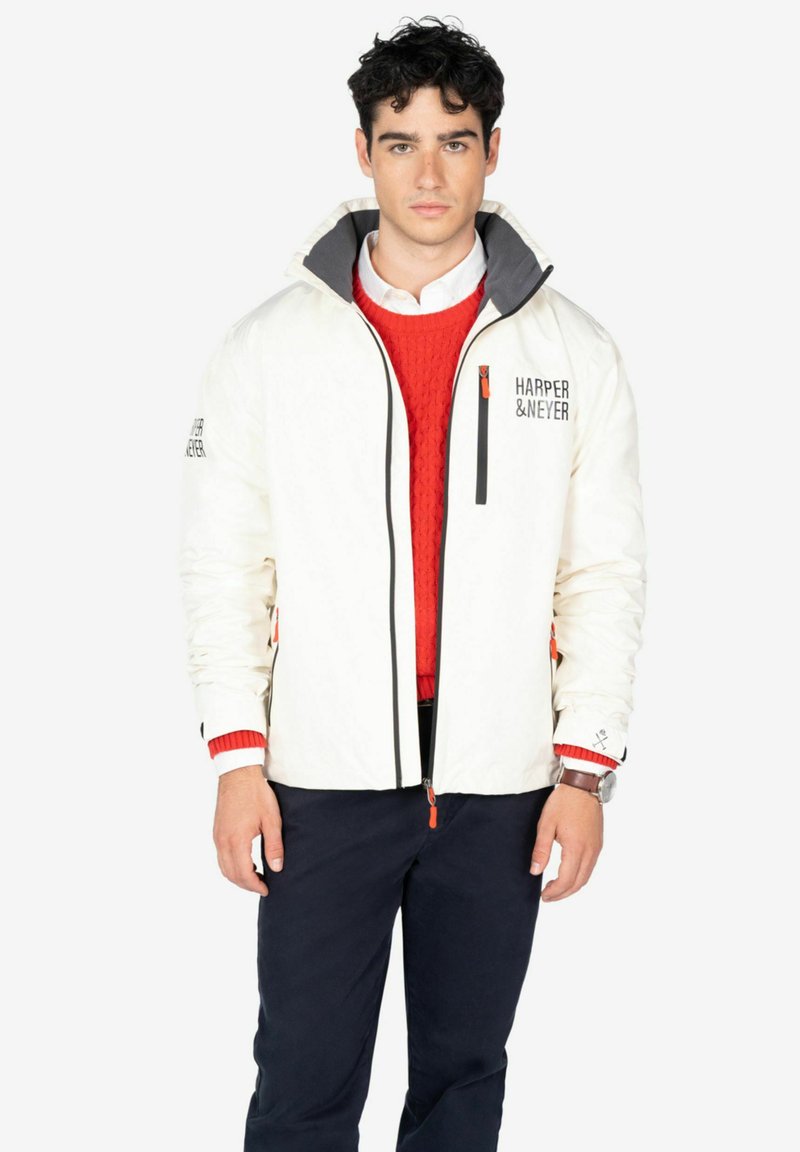 Chaqueta impermeable blanca con cuello gris, cremallera negra y detalles en naranja. Presenta un logo en el lado izquierdo del pecho y bolsillos en las mangas.