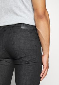Vaqueros de mezclilla negros con un acabado texturizado, que presentan una etiqueta de cuero en la cintura. El diseño del bolsillo trasero incluye un patrón de costura simple.
