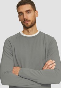 Felpa grigia a maniche lunghe con collo rotondo e finitura testurizzata, dotata di un colletto interno bianco e maniche raglan. Senza loghi o motivi visibili.