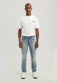 Man staat in een witte "NO STRESS CLUB" t-shirt, lichte spijkerbroek en witte sneakers op een effen lichte achtergrond.