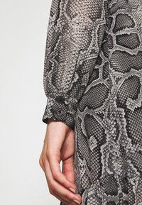 Langärmliges Kleid aus einem Stoff mit schwarz-grauem Schlangenprint, ausgestattet mit einem knöpfbaren Ärmelabschluss und strukturierten Mustern, die Schuppen ähneln.