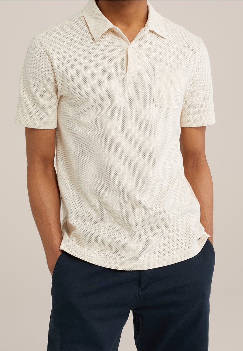 Homme portant un polo à manches courtes texturé de couleur blanc cassé avec une poche sur la poitrine et un pantalon bleu marine, les mains dans les poches.