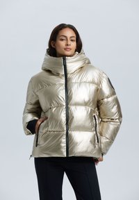 Guldmetallisk pufferjacka med hög krage, framsidans dragkedja och sidofickor. Den har en slät textur och kviltad design.