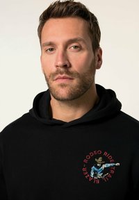 Černá mikina s kapucí, na které je vyšitý kruhový grafický motiv kovboje s červeným textem "BLAZER-RODEO RIDE TRAIL." Měkká tkanina s volným střihem.