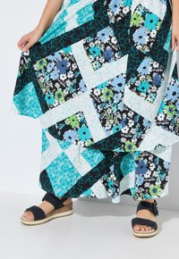 Abito lungo floreale in un design patchwork con colori blu, verde e nero. Una persona indossa sandali con zeppa blu navy con una cinghia incrociata.