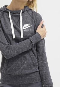 Mörkgrå hoodie tillverkad av mjukt, strukturerat tyg. Har vita dragsnören, en ficka fram och en logotyp på bröstet.