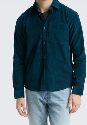 Homme portant une chemise en velours côtelé vert foncé à manches longues par-dessus un t-shirt noir avec un jean bleu clair, debout, les bras détendus le long du corps.