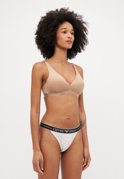 Soutien-gorge sans fil beige avec un décolleté en V profond et des bretelles larges ; associé à un string blanc doté d'une ceinture de marque noire. Texture lisse.