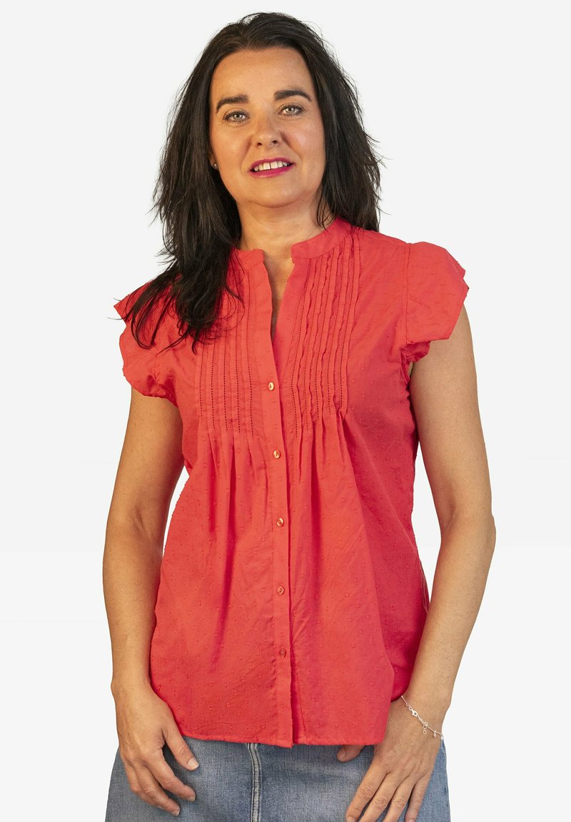 Blusa de color coral con mangas cortas con volantes, pliegues en la parte delantera y cierre de botones. Fabricada con un tejido ligero con una textura sutil.