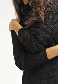 Maglione nero lavorato a maglia caratterizzato da un design ampio e testurizzato con maniche larghe e un collo alto. Il materiale sembra morbido e spesso.