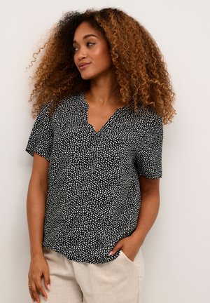 Blouse noire à manches courtes avec motif abstrait blanc, décolleté en V, tissu léger, coupe décontractée, associée à un pantalon beige.