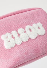 Vaipan vaaleanpunainen pussi, jossa on pörröiset valkoiset kirjaimet, jotka sanovat "BISOU". Pussissa on teksturoitu, pehmeä pinta ja hillitty raidallinen kuvio.