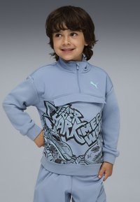 Ragazzo che sorride e guarda di lato, indossando un maglione Puma azzurro chiaro con testo in stile graffiti e pantaloni azzurri chiari.