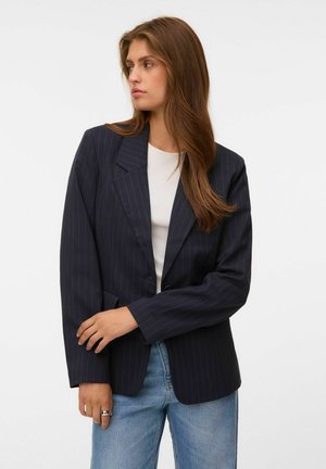 Kvinde med langt brunt hår iført en mørk koksgrå blazer med striber over en hvid skjorte og blå jeans, der kigger til siden med krydsede arme.
