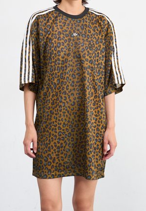 Løs t-shirt-kjole med leopardprint, sort krave og hvide tre-stribede detaljer på ærmerne, båret af en stående person.