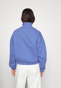 Suéter de forro polar azul con cuello alto, dobladillo elástico y mangas holgadas, combinado con pantalones amplios de color claro.