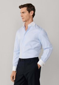 Mężczyzna w jasnoniebieskiej koszuli z białym kołnierzykiem i mankietami, ciemnych spodniach, stojący z jedną ręką w kieszeni, patrzący w lewo.