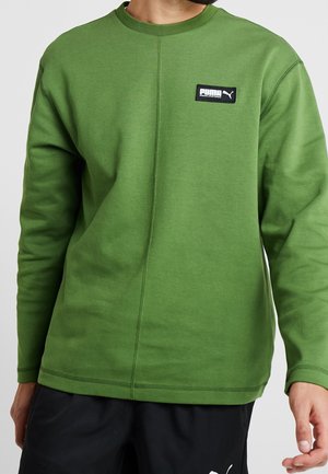 Mand iført grøn Puma langærmet sweatshirt med logopatch på brystet og sorte bukser med Puma-logo.