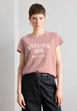 Osoba s krátkymi vlasmi, ktorá nosí červeno-biele pruhované tričko s textom "ATELIER 2010 MOS MOSH" a tmavé nohavice pred jednoduchou stenou.