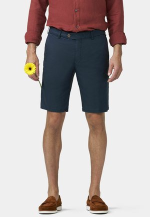Mann trägt marineblaue Shorts, rostfarbenes Langarmshirt, braune Loafers, hält eine gelbe Blume in der linken Hand vor einem einfarbigen Hintergrund.