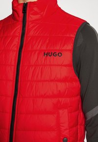 Rote gepolsterte Weste mit hohem Kragen, ausgestattet mit einem schwarzen Reißverschluss und Logo. Strukturierter Stoff mit horizontalen Nähten, getragen über einem schwarzen Langarmshirt.