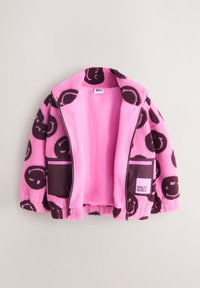 Veste polaire rose avec des motifs de visages souriants foncés, fermeture éclair à l'avant, deux poches foncées et étiquette "SMILEY WORLD" sur la poche droite.