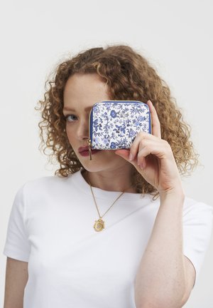 Floral-patterned zippered wallet in white and blue, featuring a gold accent. Held in a hand against a neutral background.